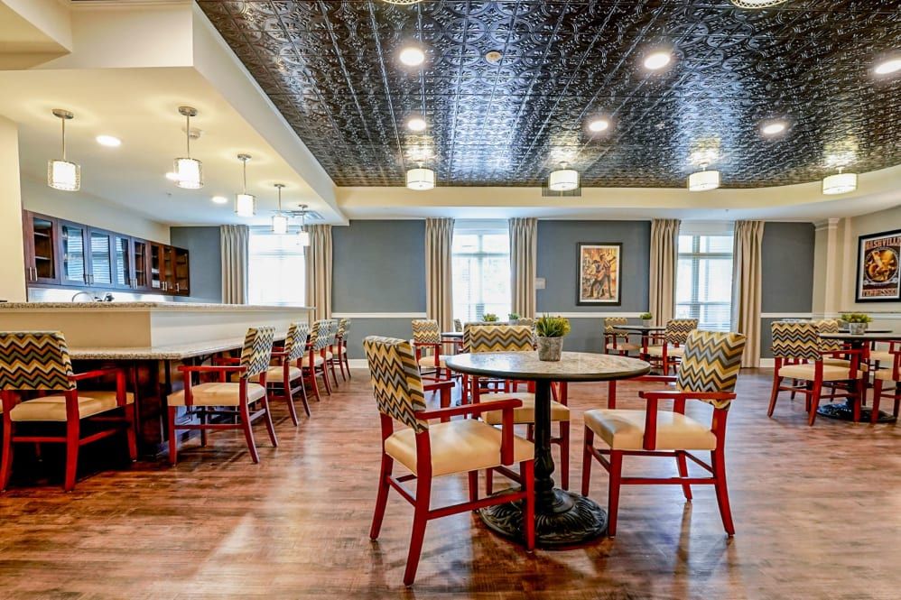 A spacious dining area with round tables and patterned chairs, featuring a decorative ceiling and wooden flooring.