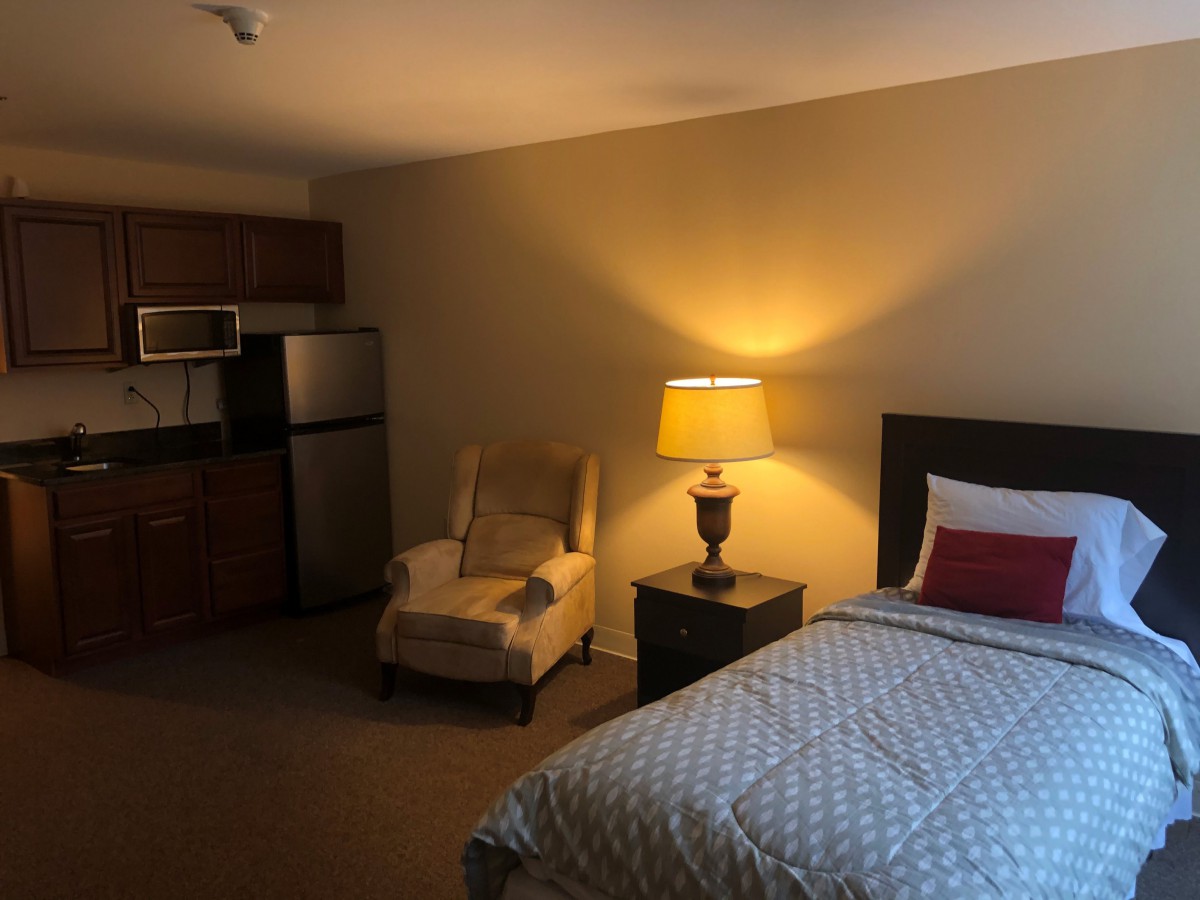A cozy hotel room featuring a bed, nightstand with a lamp, chair, and kitchenette.