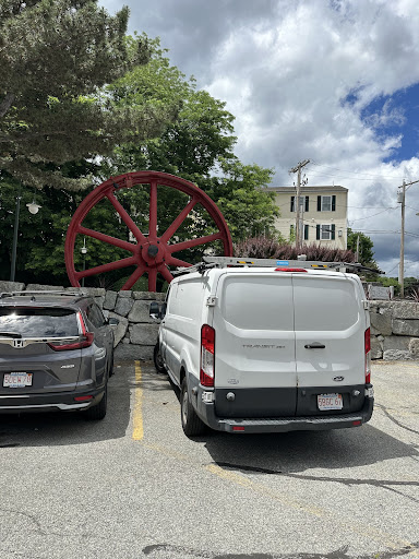 A parked white van in front of a large red historic wheel, with a backdrop of a cloudy sky and a building.