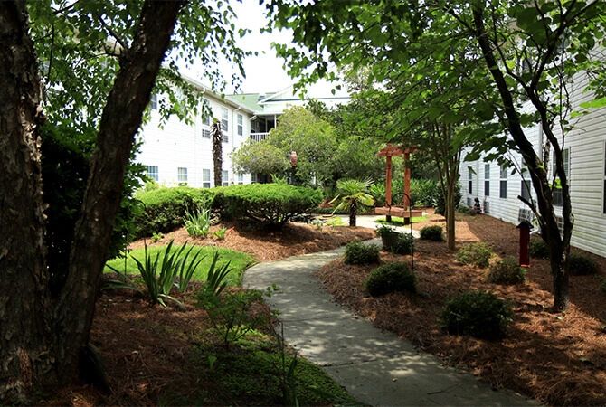 A serene garden with a winding pathway surrounded by lush greenery and benches, adjacent to a residential building.