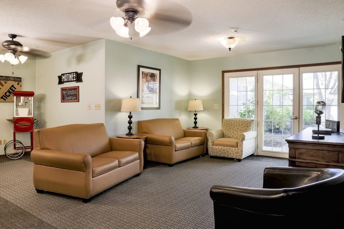 A cozy living room with a ceiling fan, sofas, chairs, lamps, and a view of the outdoors through large windows.