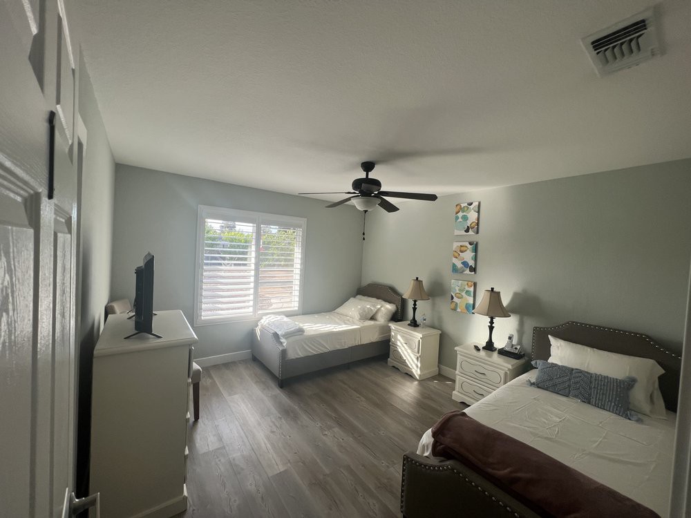 A modern bedroom featuring two twin beds, a ceiling fan, and ample natural light filtering through the windows.