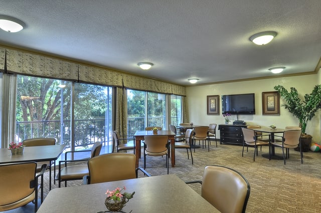 A spacious dining room with multiple tables and chairs, large windows, and a wall-mounted television.