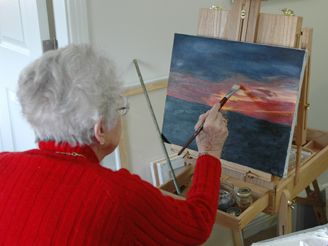 An elderly woman painting a landscape at an easel.