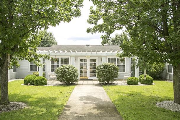 A charming white house with large windows surrounded by a lush garden and trees, featuring a pathway leading to the entrance.