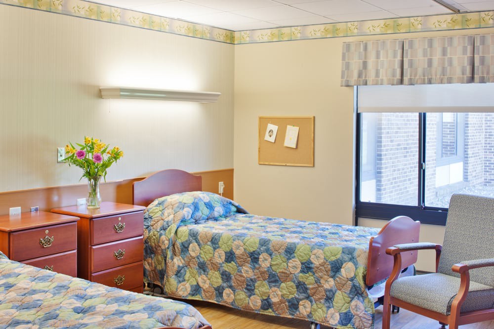 A hospital room with two twin beds, a wooden dresser with flowers, and a window with blinds.