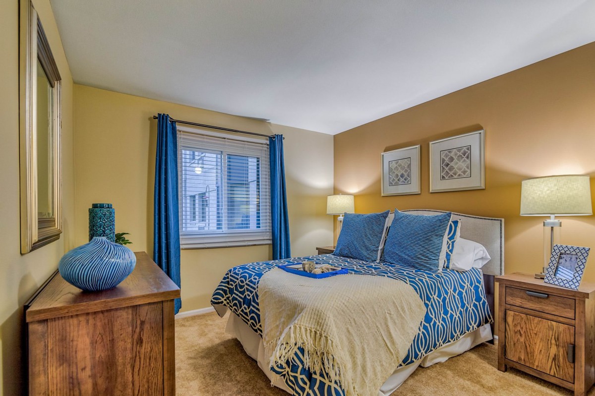 A cozy bedroom with blue and beige decor, featuring a comfortable bed, a lamp, and beautiful artwork on the walls.