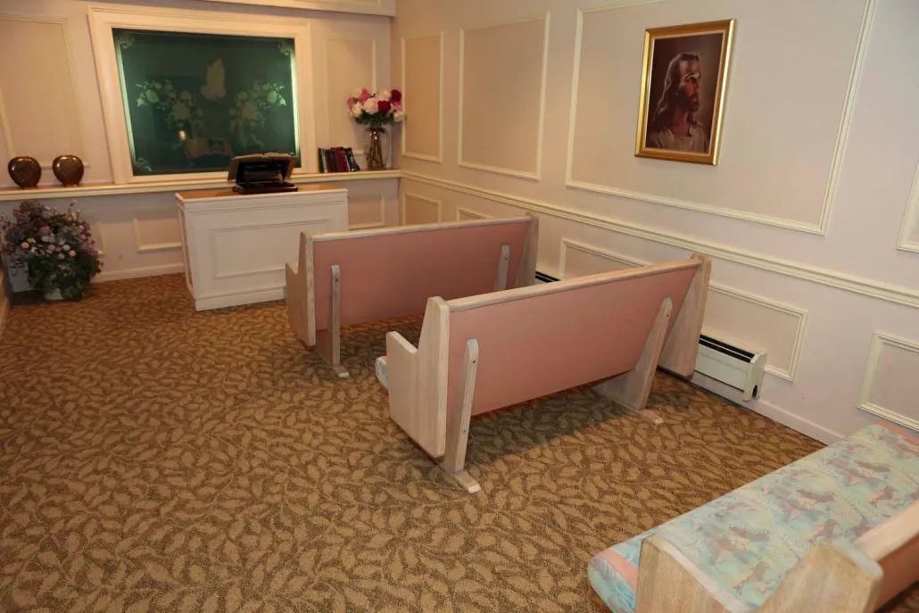 A small, carpeted room with two pews facing a podium and a window, adorned with flowers and a painting on the wall.