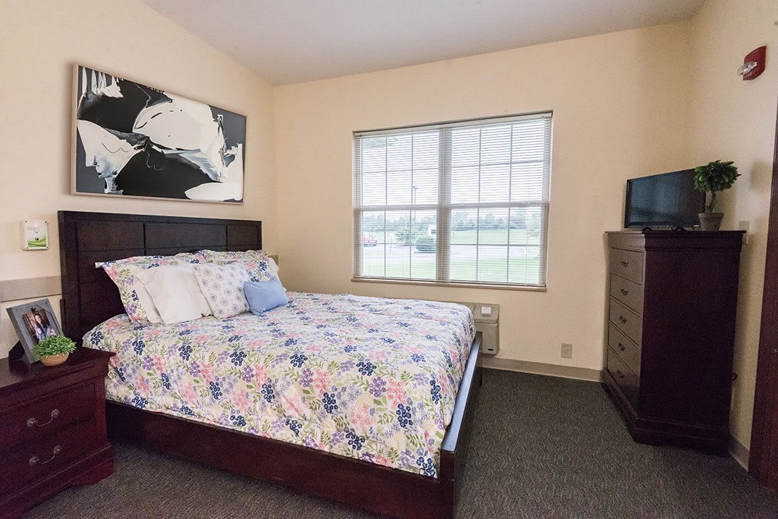 A cozy bedroom with a large window, a bed with floral bedding, and a dresser.