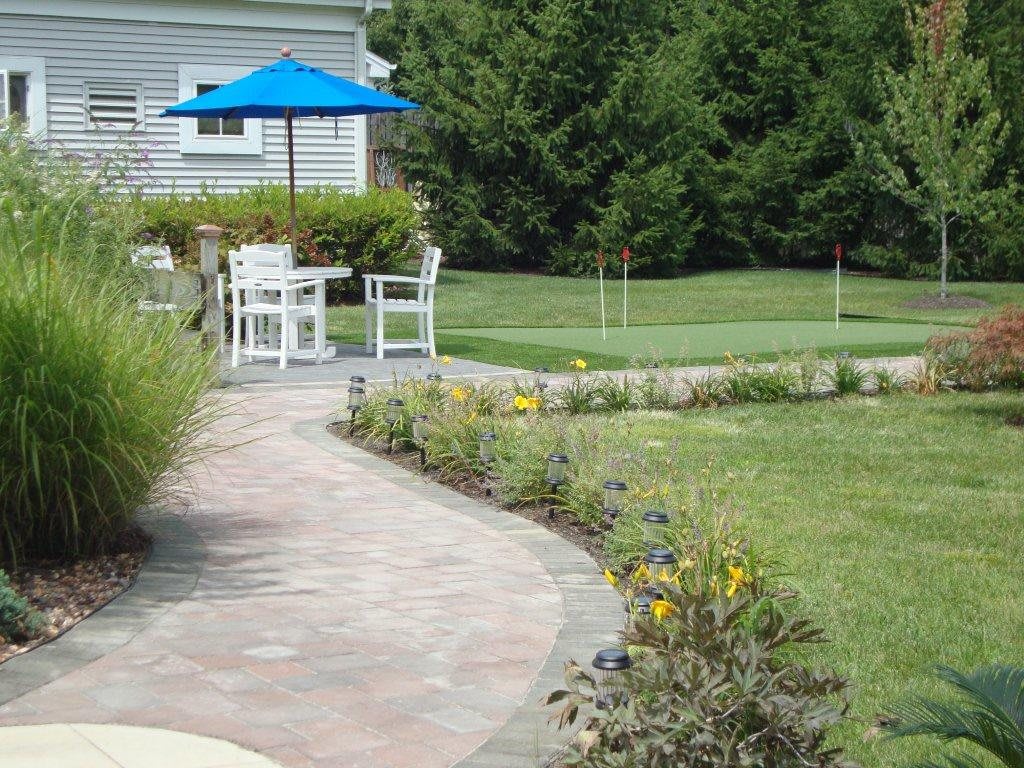 A tranquil garden with a paved pathway leading to a patio area featuring white furniture and a blue umbrella amidst lush greenery.