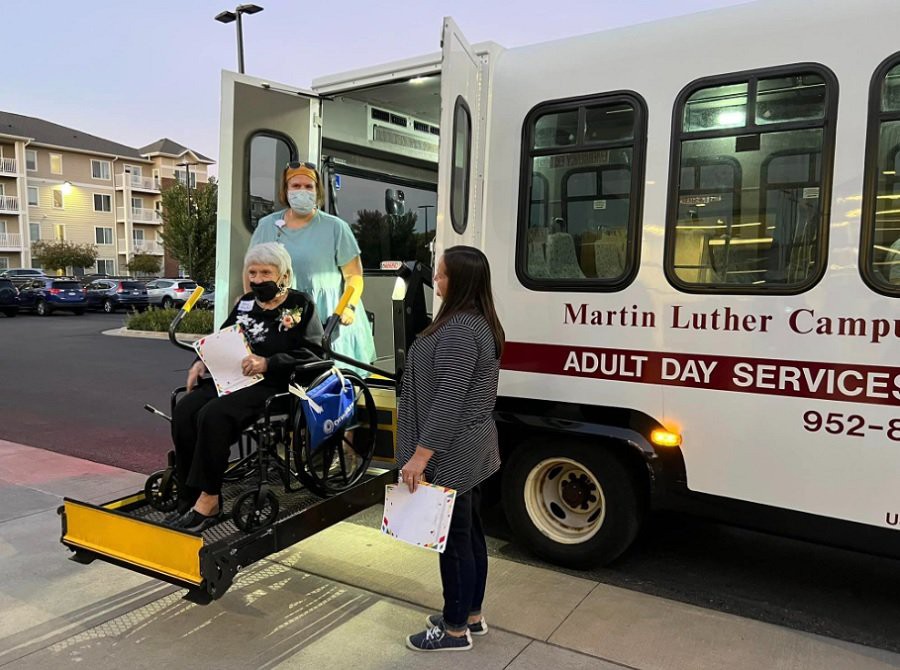 A healthcare worker assists a person in a wheelchair onto an adult day services bus.