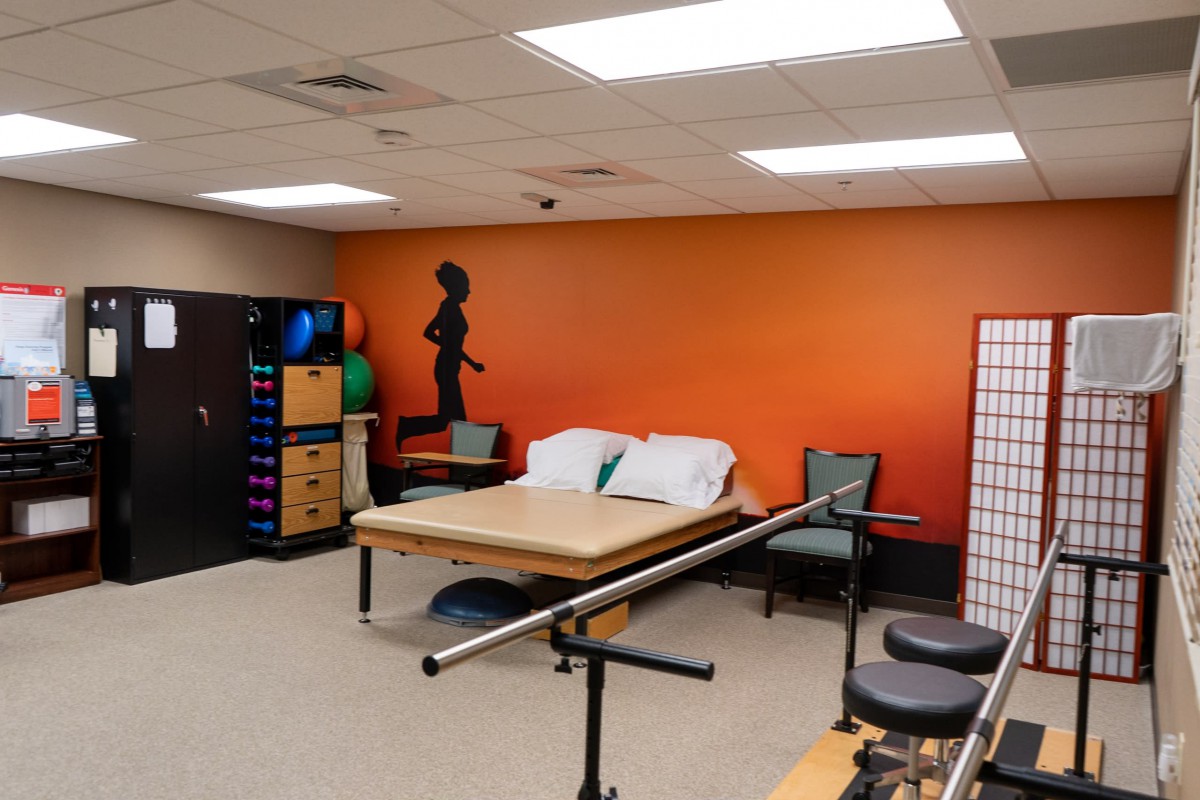 A rehabilitation room equipped with a treatment bed, exercise equipment, and parallel bars against an orange accent wall.