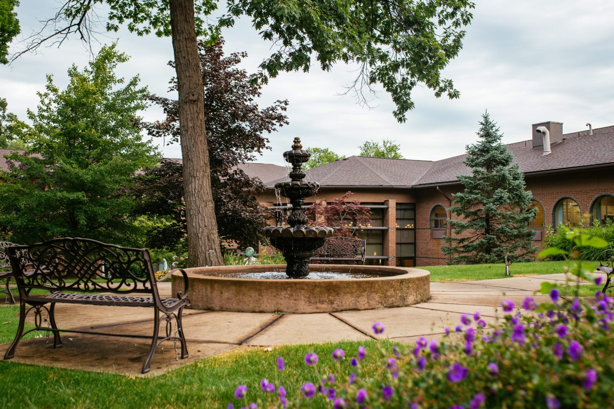 A tranquil garden scene with a central fountain, surrounded by trees and blooming flowers.