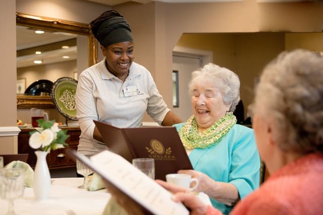 A caregiver assisting an elderly woman with a menu at a senior living facility.