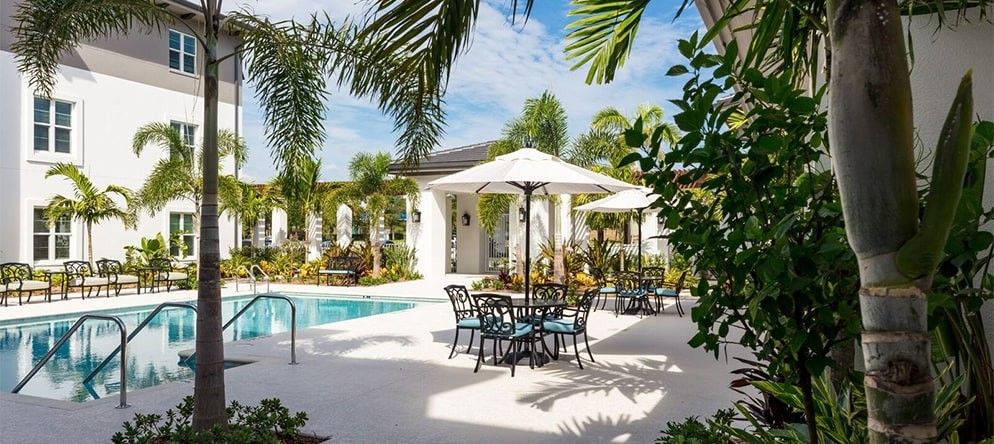 A sunny patio area with a pool, surrounded by tropical palm trees and furnished with tables, chairs, and umbrellas.