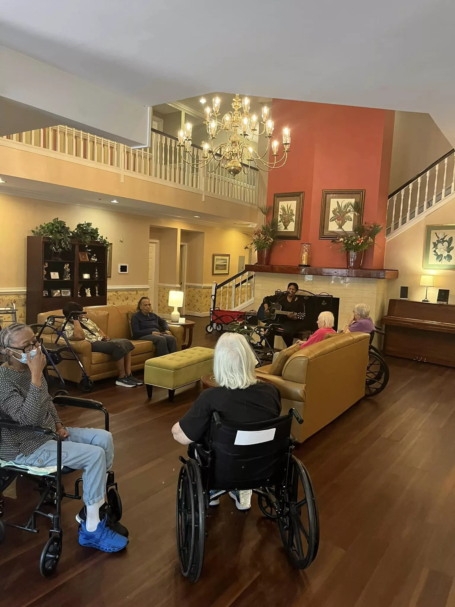 A cozy nursing home lounge where residents are gathered, some in wheelchairs, enjoying a communal space with sofas and a chandelier.
