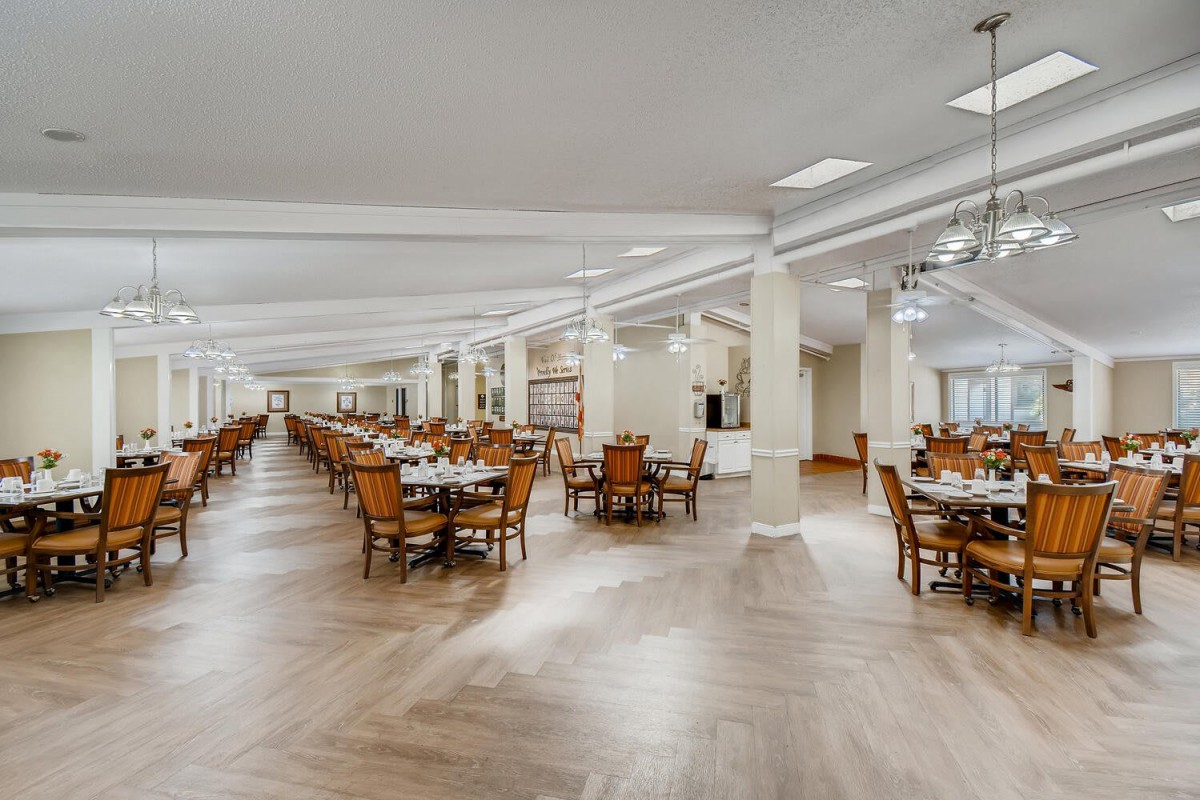 A spacious dining hall with wooden flooring, filled with neatly arranged tables and chairs.