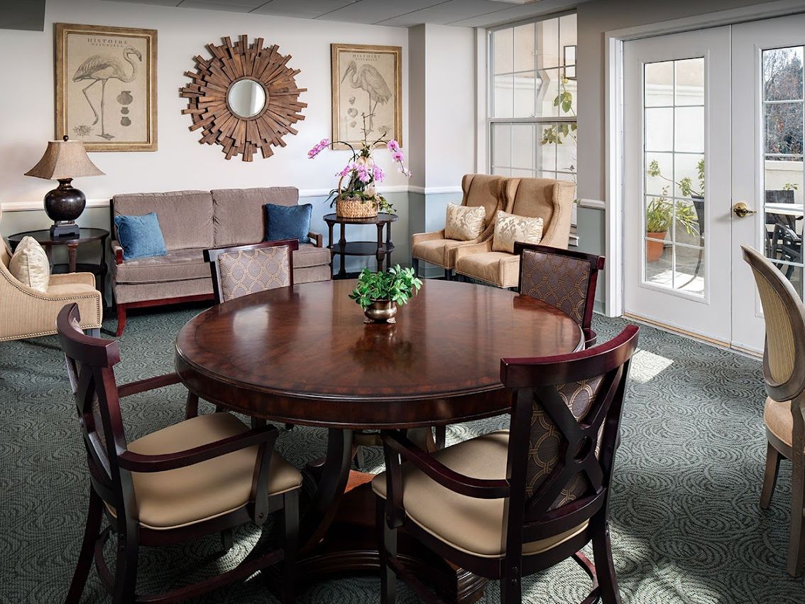 A cozy living room featuring a round wooden table surrounded by chairs, with a couch and armchairs near the window.