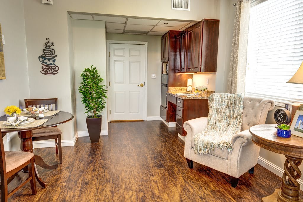 A cozy living room with wood flooring, featuring an armchair, potted plant, and compact kitchen area.