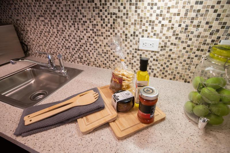 A kitchen countertop with cooking essentials including wooden utensils, pasta, olive oil, and canned food against a mosaic-tiled backsplash.