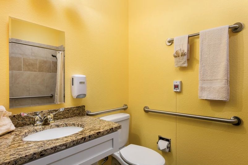 A well-lit bathroom with yellow walls and a granite countertop, equipped with a sink, toilet, mirror, towels, and handrails.