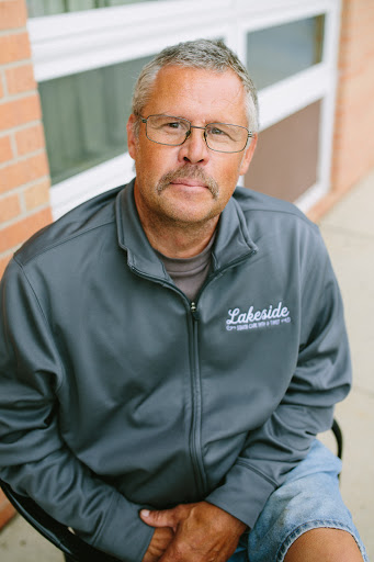 A thoughtful man seated outdoors, wearing a gray jacket and glasses.