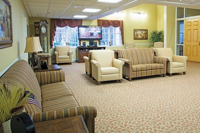A cozy living room with multiple sofas and chairs arranged around a television, featuring carpeted flooring and large windows.