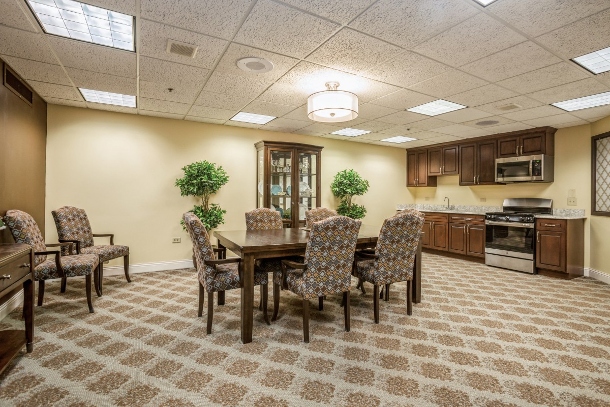 A well-lit dining room with a table and chairs, surrounded by plants and a kitchen area.
