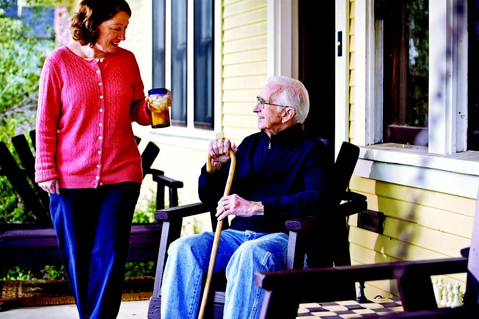 An elderly man sitting on a porch, holding a walking cane, while conversing with a woman standing nearby.