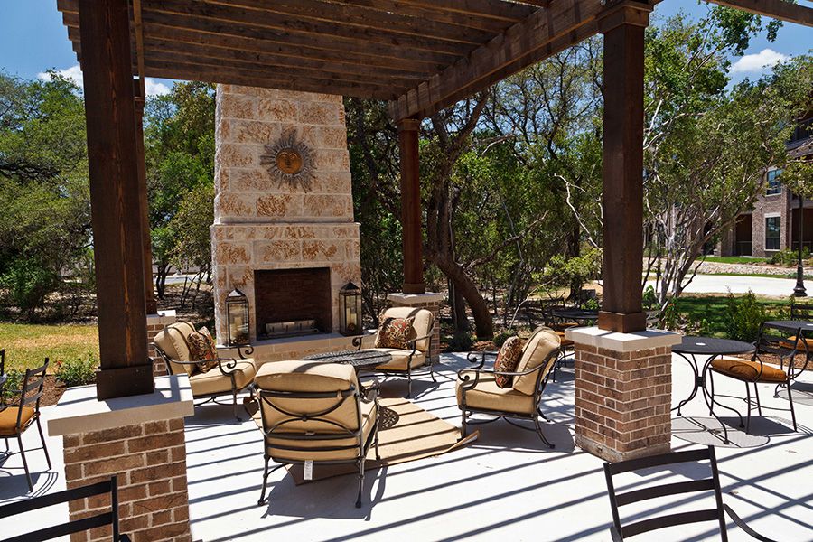 An outdoor patio with comfortable seating around a fireplace under a pergola, surrounded by trees.