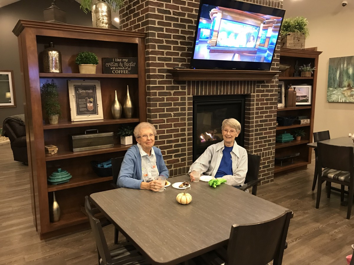 Two elderly individuals sitting at a table in a cozy room with a TV and fireplace.