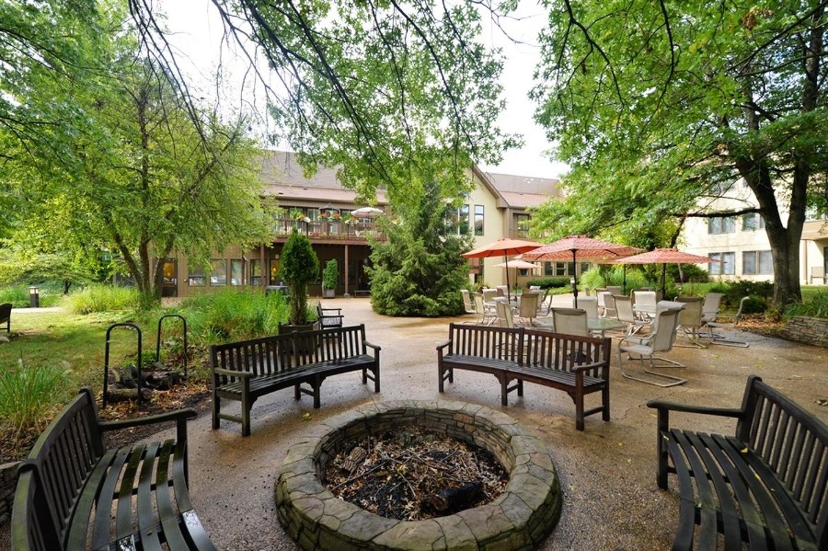 A serene courtyard featuring wooden benches surrounding a stone fire pit, with lush greenery and outdoor seating areas.