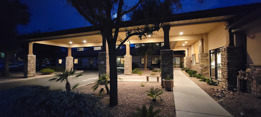 A welcoming entrance to a building illuminated at night, surrounded by palm trees and decorative landscaping.