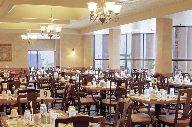 An empty dining hall with neatly arranged tables and chairs, illuminated by chandeliers.