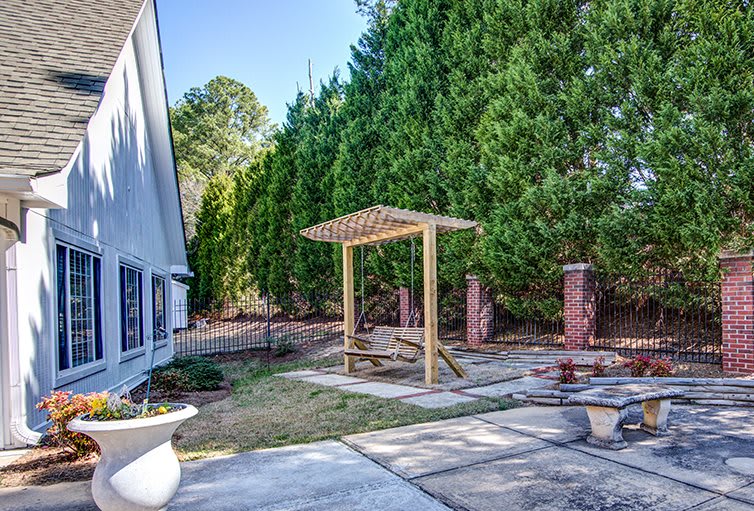 A backyard view featuring a small wooden pergola, surrounded by lush green trees and a paved patio area.