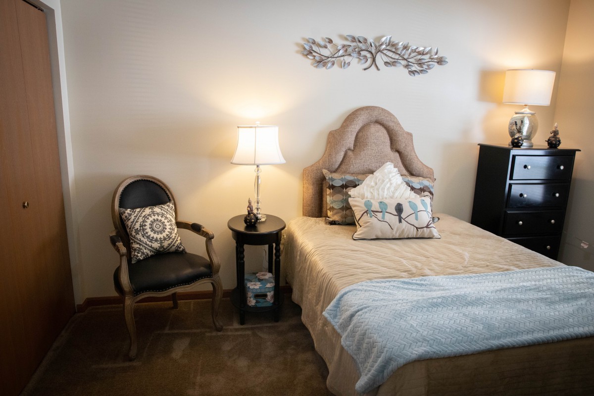 A cozy bedroom with a classic design featuring a bed, nightstand with a lamp, and an elegant decorative chair.