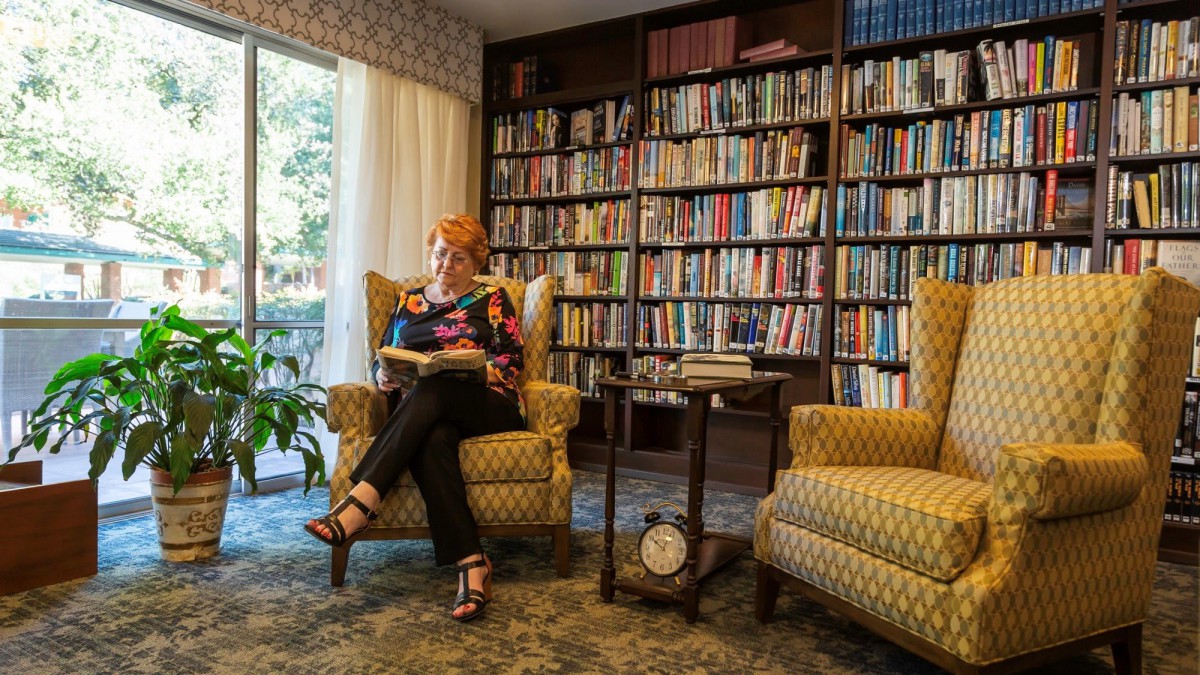 A woman sitting in an armchair reading a book in a cozy library room.