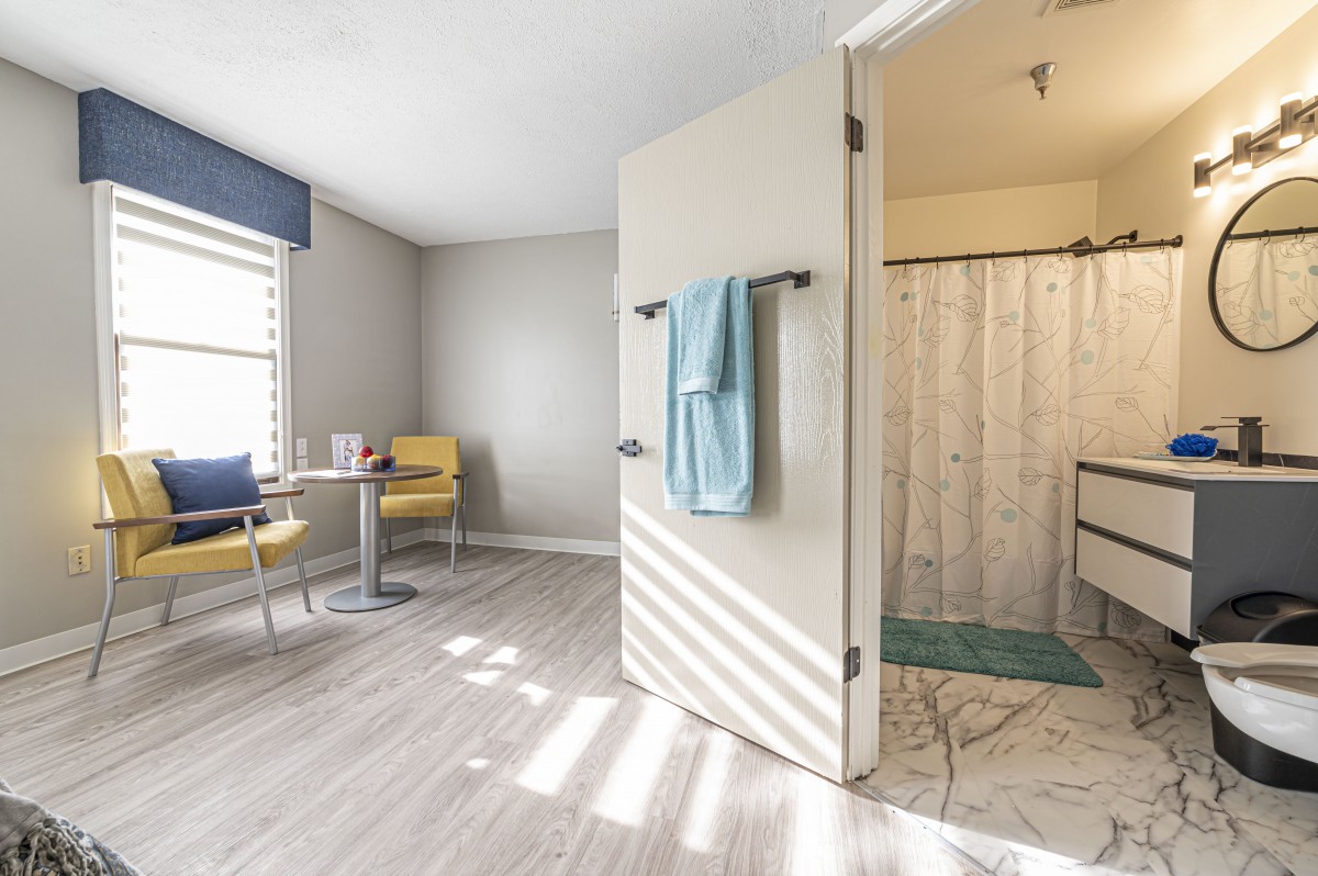 A modern interior space with a view into a bathroom, featuring light wood flooring, a small table with chairs, and a towel hanging on the door.