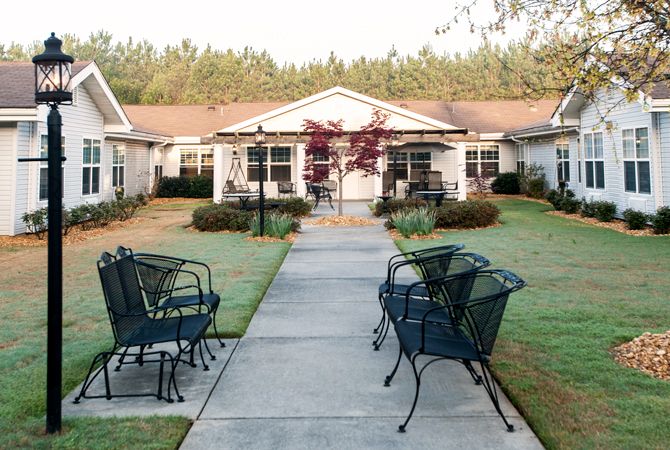 A serene outdoor scene with a pathway leading to houses, flanked by chairs and a well-maintained garden.