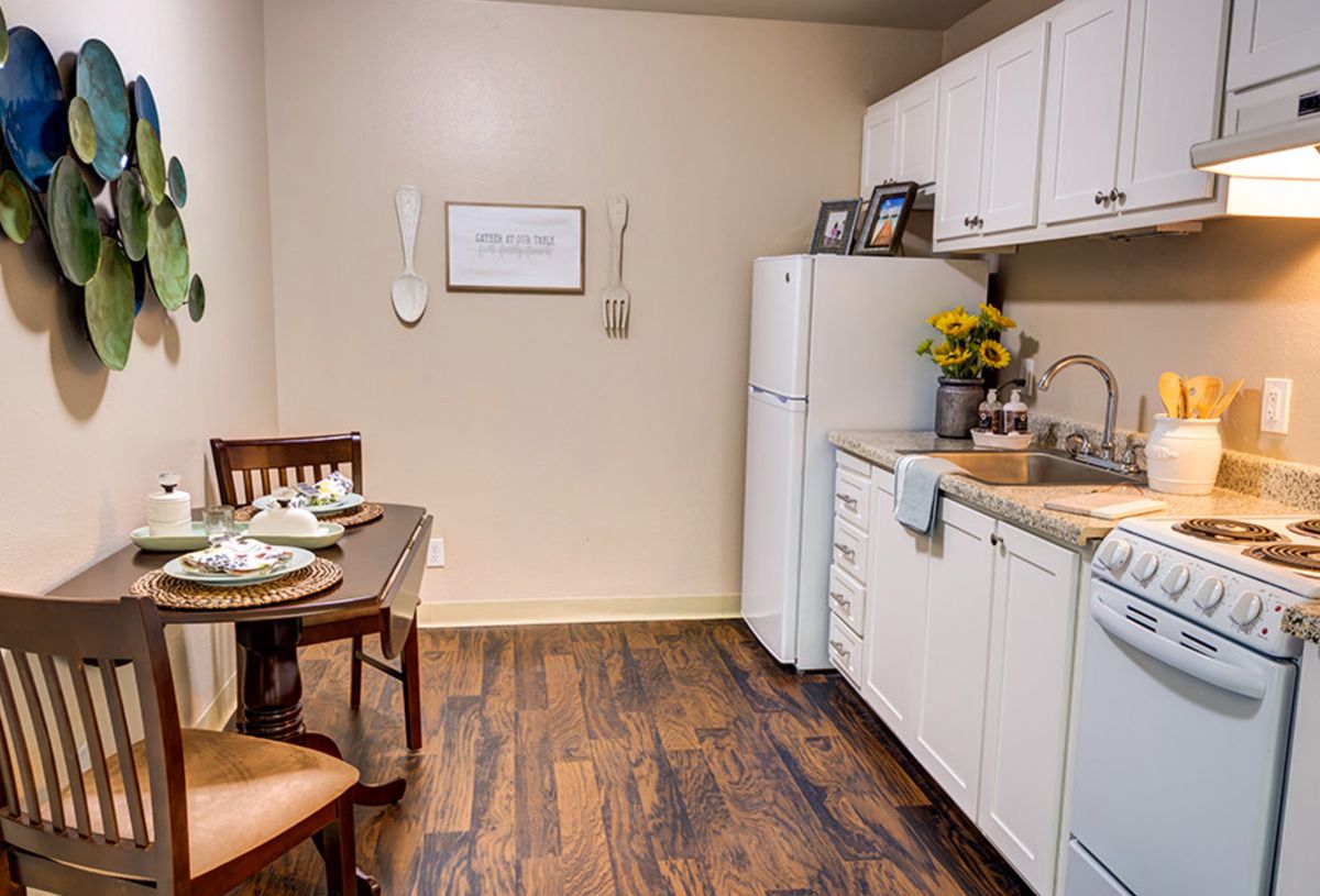 A small kitchen and dining area with wooden flooring, white cabinets, a refrigerator, and a table set for two.