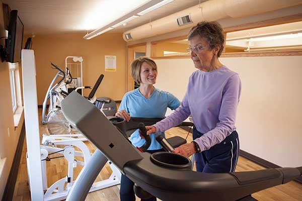 An elderly woman exercises on a treadmill with the assistance of a trainer in a gym setting.