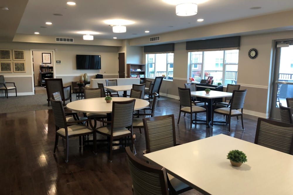 A community dining area with several tables and chairs, featuring wooden floors and large windows.