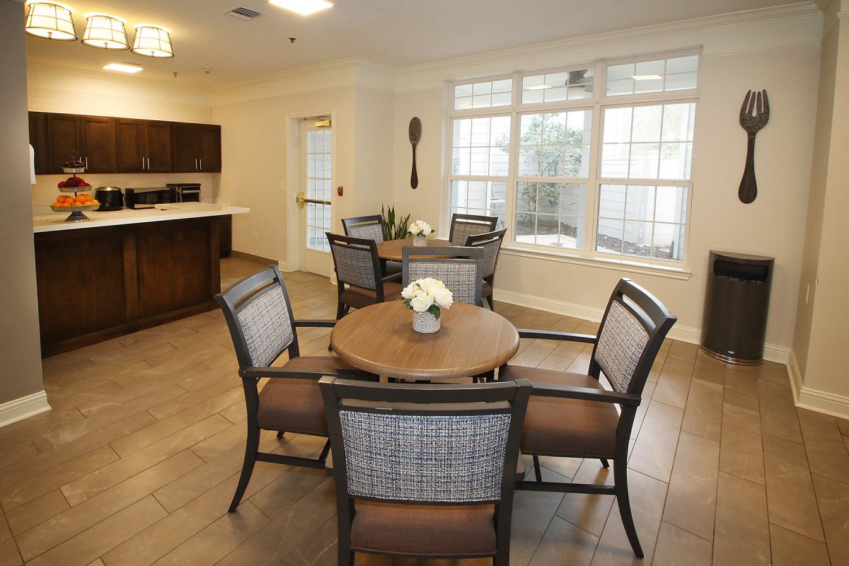 A cozy dining area with round tables and chairs, adjacent to a kitchen, featuring large windows that allow natural light to fill the space.