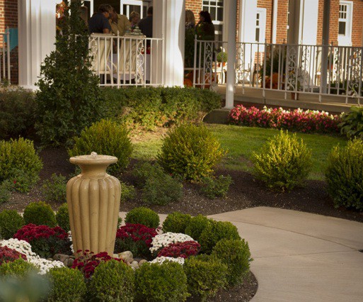 A meticulously landscaped garden with a central urn surrounded by vibrant flowers and neatly trimmed shrubs adjacent to a patio.