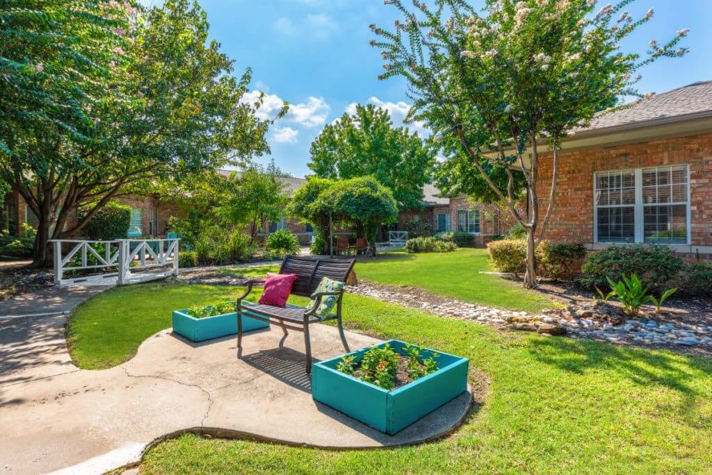 A peaceful garden with a bench surrounded by lush greenery and vibrant planters under a sunny sky.