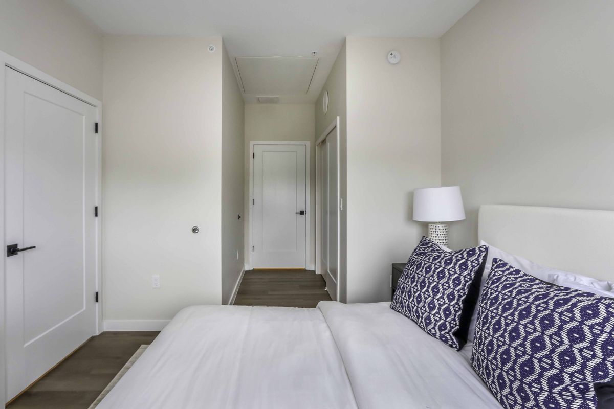 A minimalist bedroom with white walls, a neatly made bed, patterned pillows, and a bedside lamp.