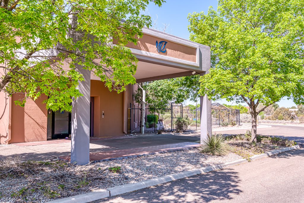 A modern building entrance shaded by a canopy, surrounded by green trees on a sunny day.
