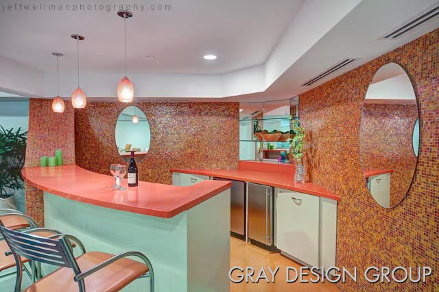 A stylish retro-themed kitchen bar area featuring orange tile walls, a pink countertop, modern pendant lights, and a bottle of wine on the counter.
