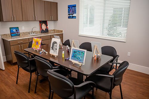 An office setting with an art studio, featuring several paintings on easels displayed around a table surrounded by chairs.