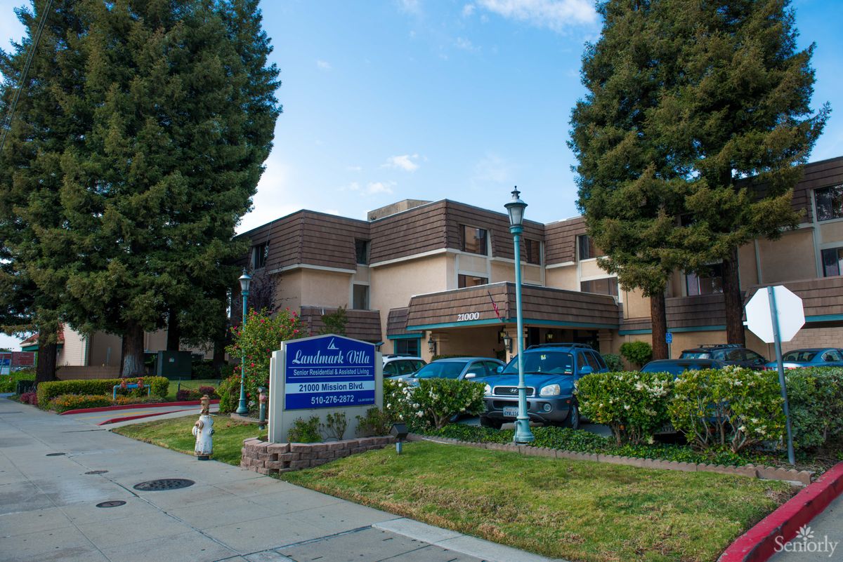 A residential building with a sign and parked cars, surrounded by trees under a cloudy sky.